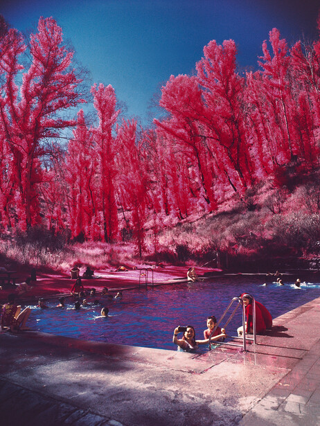 Infrared landscape with magenta trees surrounding a blue lake and sandy beach with people.