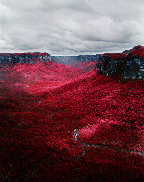 Infrared landscape photograph showing deep red vegetation covering rolling hills beneath a cloudy sky.