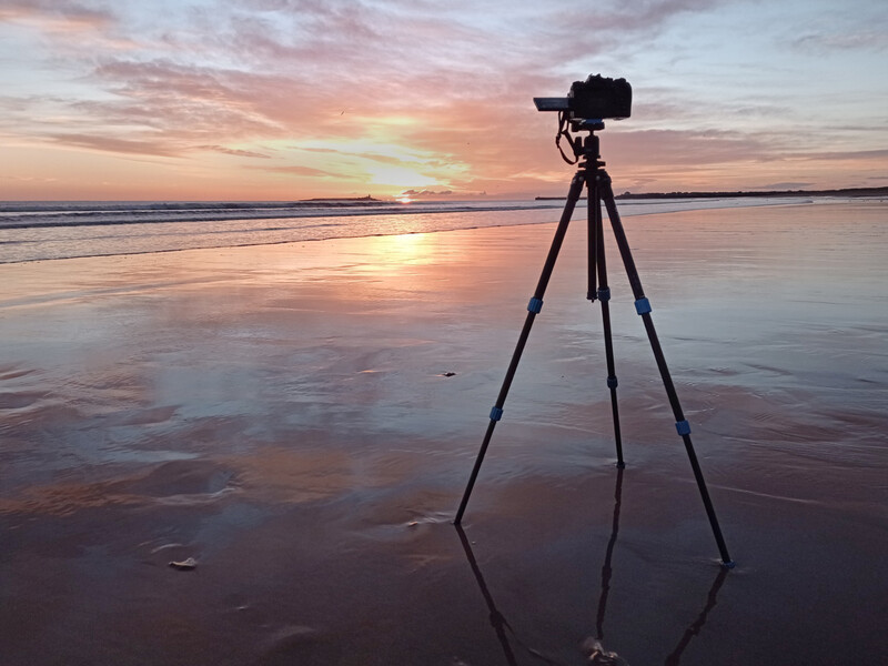 Camera mounted on tripod on wet beach sand during golden hour sunset.
