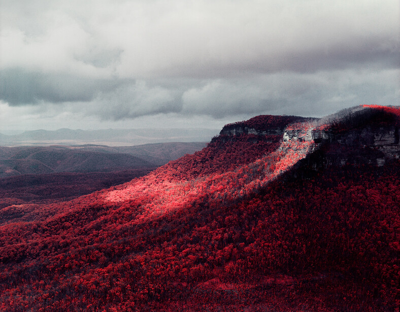 Mountain ridge covered in vibrant red autumn foliage beneath dramatic cloudy sky.