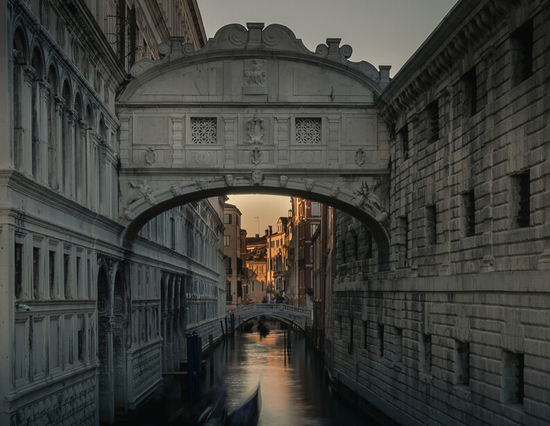 Venetian canal framed by the Bridge of Sighs, with warm-lit buildings visible through the archway and water reflections below.