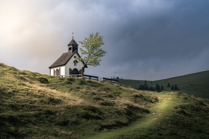 Small white chapel with dark roof perched on rolling green hillside under dramatic cloudy sky.