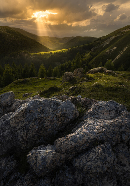 Sunlit valley framed by forested mountains with rocky boulders in foreground.