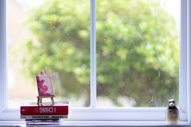 Miniature easel with pink painting positioned on a windowsill next to stacked books and a small figurine, with bright green foliage visible outside the window.