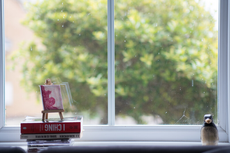 Windowsill still life with stacked books, small canvas, and ceramic figurine against blurred green foliage.