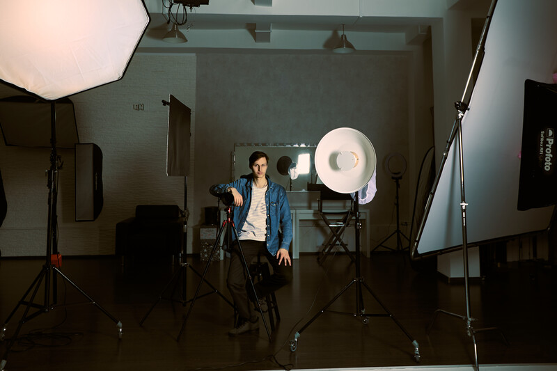 Portrait photographer posing under studio lights in a professional shooting setup with multiple softboxes and reflectors.