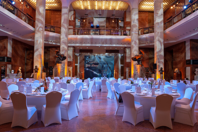 Large ballroom with round tables draped in white linens, ornate columns, and warm amber ceiling lighting.