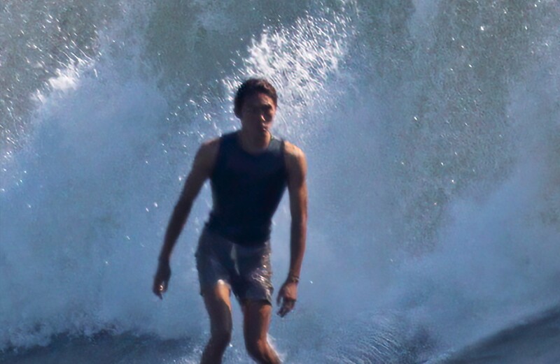 Surfer standing in shallow water as a large wave crashes overhead, backlit by bright sunlight filtering through the water.