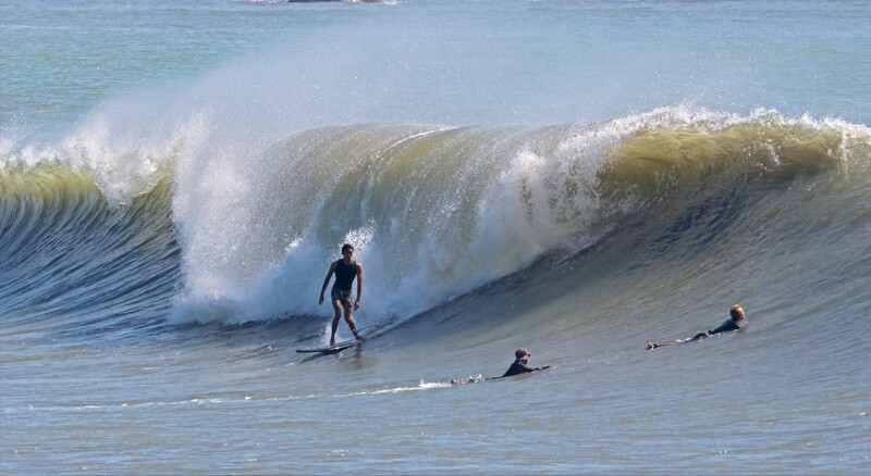 Surfer standing on board in shallow water facing a massive breaking wave with several swimmers nearby.