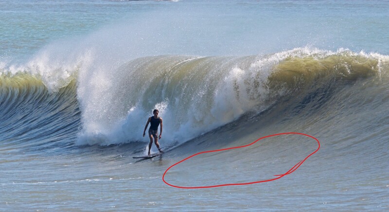 Surfer in black wetsuit standing in shallow water facing an enormous wave with white foam crest.