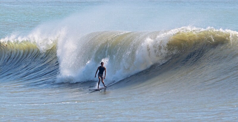 Surfer riding a large wave in shallow water, captured with bright natural daylight and clear visibility of wave texture and spray.