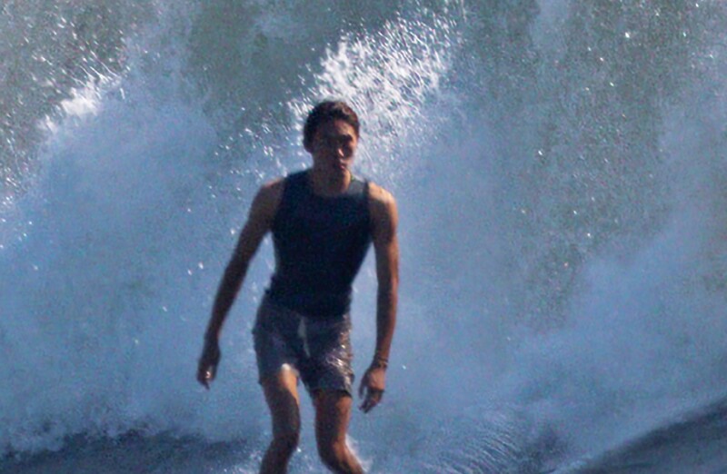 Surfer standing in shallow water as a large wave crashes over him with dramatic spray and backlighting.