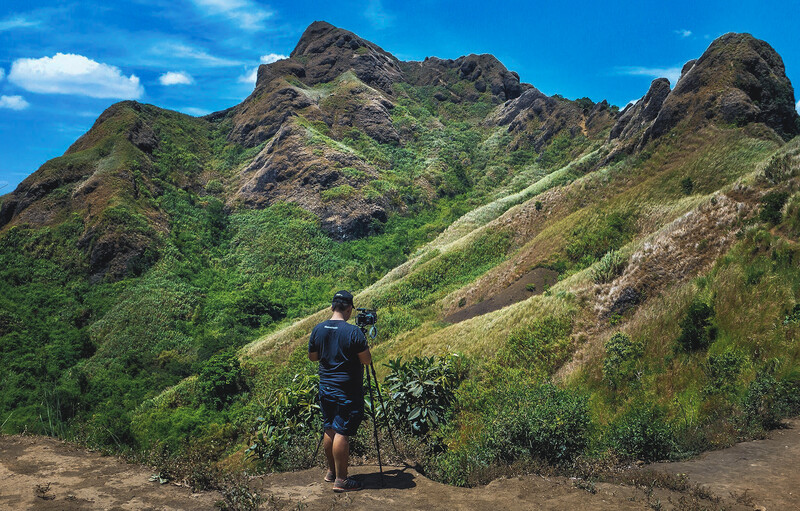 Hiker with trekking poles standing on a trail facing a lush, verdant mountain with dramatic rocky peaks.