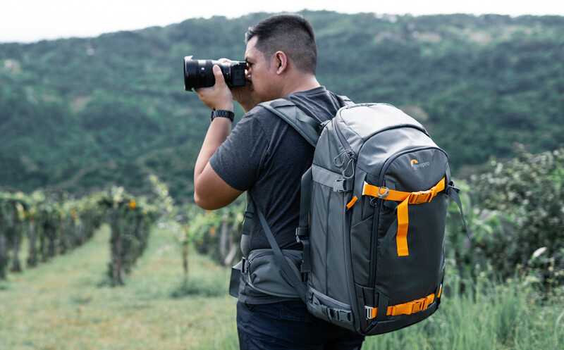 Photographer with backpack looking through binoculars in a forested hillside landscape.