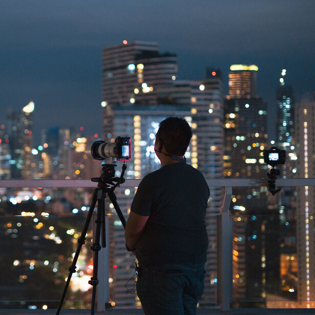 Photographer silhouetted against illuminated city skyline at twilight, operating camera on tripod.