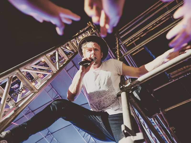 Performer singing into microphone on stage with dramatic purple lighting and hands raised above.