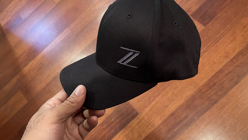 Black baseball cap with stylized Z logo held in hand against wooden floor background.