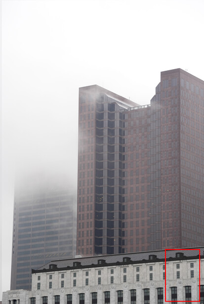 Tall brick buildings shrouded in dense fog with a lower multi-story structure in the foreground.