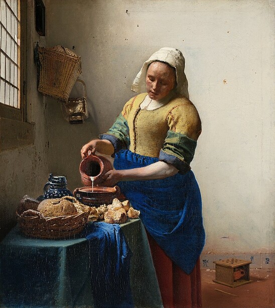 Woman in yellow and blue dress pouring milk into a bowl of bread on a table.