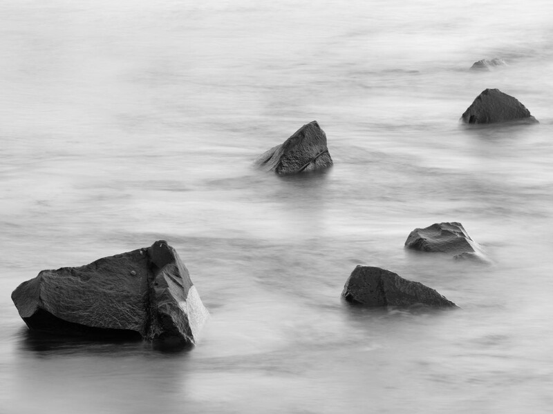 Dark rocks protruding from misty ocean water with a smooth, ethereal appearance.
