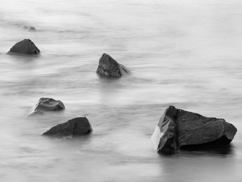 Dark rocks emerging from smooth, blurred water in a black and white seascape.