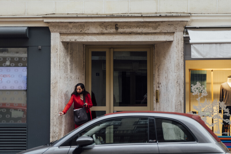 Woman in red jacket standing in doorway of weathered storefront with parked car in foreground.