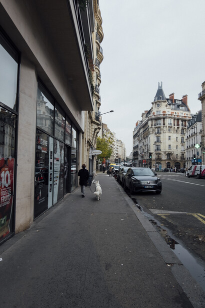 Parisian street with classical Haussmann buildings, sidewalk cafés, and parked cars under overcast sky.
