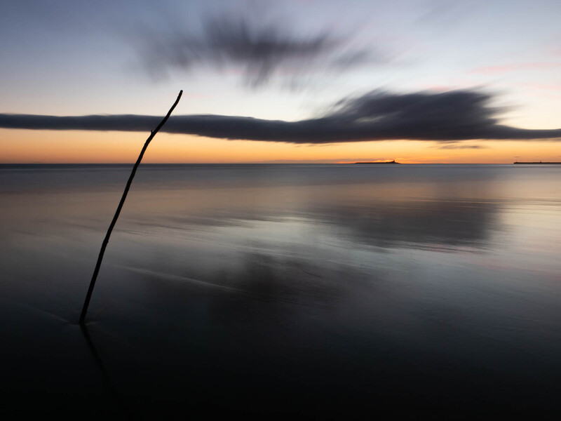 Angled wooden stick protruding from calm water at sunset with dramatic cloud formations overhead.