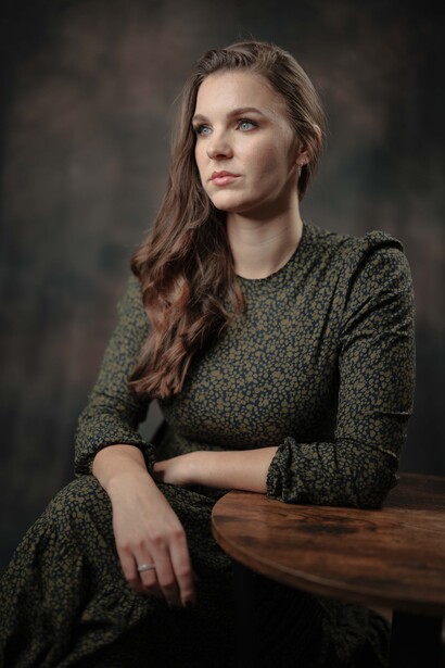 Woman with long brown hair wearing a textured green sweater, seated and gazing to the side against a dark background.