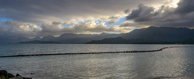 Coastal landscape with mountains across bay under dramatic cloudy sky with breaks of blue light.