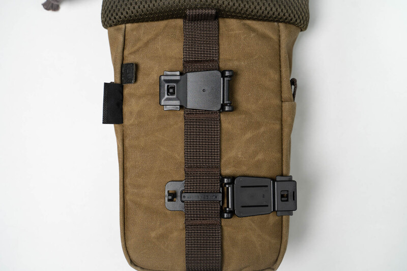 Tan canvas camera bag with brown webbing straps and dual black plastic buckle closures displayed flat.