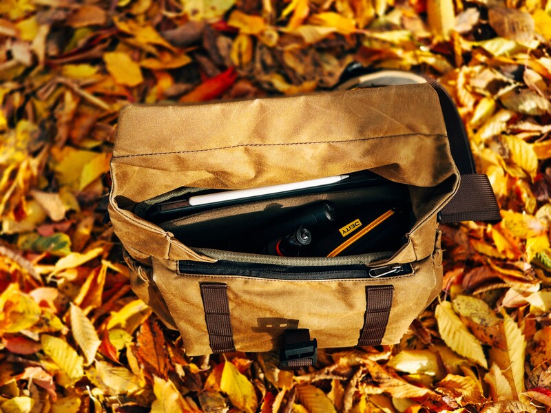 Tan canvas messenger bag open on autumn leaves, displaying camera and accessories inside.