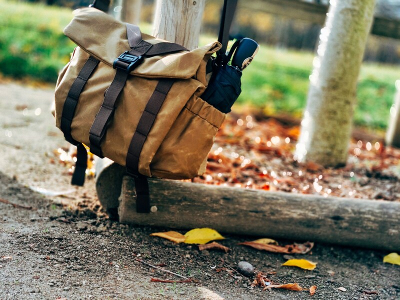 Tan canvas backpack with black straps secured to a weathered log on a forest path.