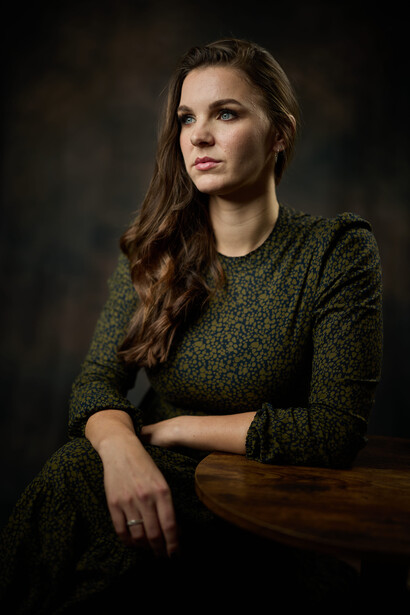 Woman in olive green sweater seated against dark background, looking toward camera.