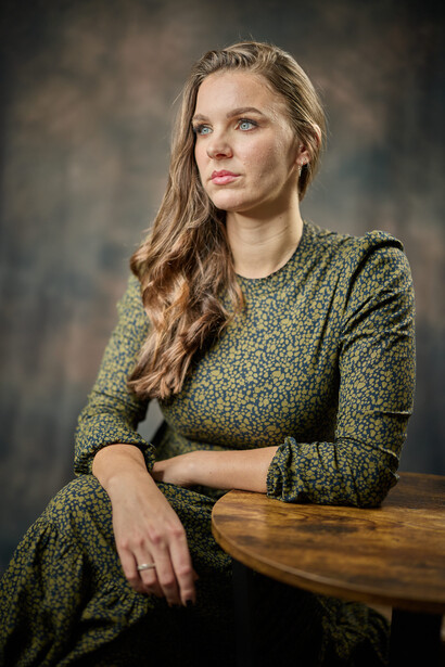 Woman in olive knit sweater leaning against wooden table with soft studio lighting.