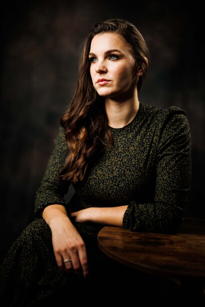 Woman with long dark hair wearing a textured dark sweater, posed against a dark background with her arm resting on wood.