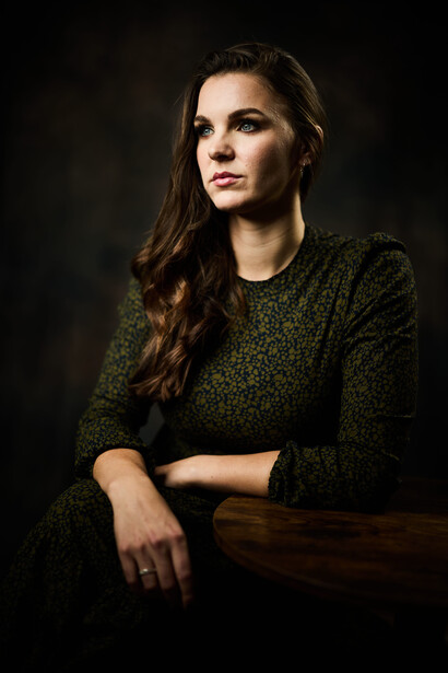 Woman with long brown hair wearing a textured green sweater, posed against a dark background with dramatic side lighting.