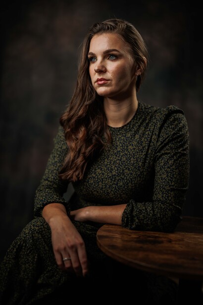 Woman in dark green knit sweater seated at wooden table against dark background.