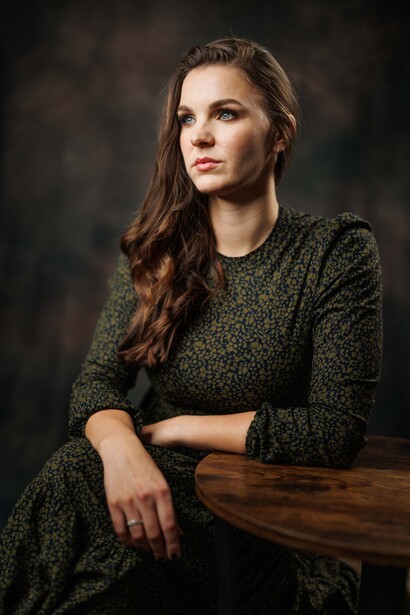 Woman in olive knit sweater seated at wooden table against dark background.