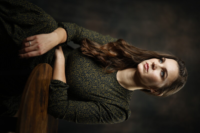 Woman with long brown hair wearing a dark patterned shirt, posed against a neutral dark background.