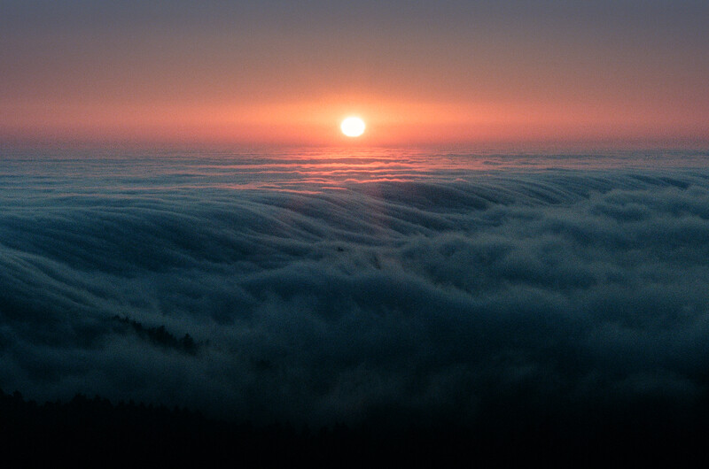 Sun rising above a sea of clouds with warm orange and pink hues lighting the sky.