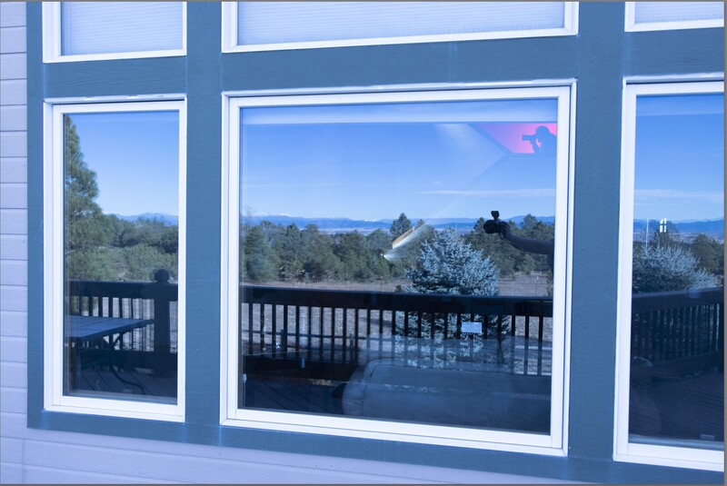 Large multi-pane window with white frames overlooking a forested valley and distant mountains under clear blue sky.