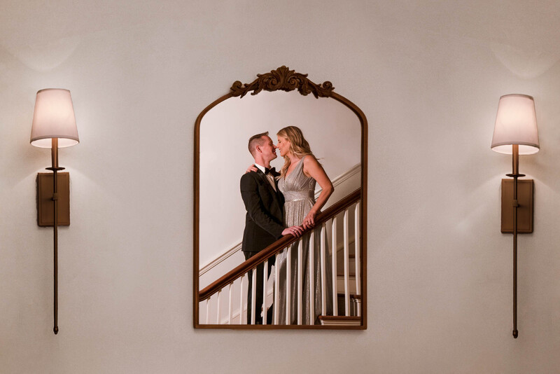 Couple kissing on a staircase, reflected in an ornate arched mirror with wall sconces on either side.