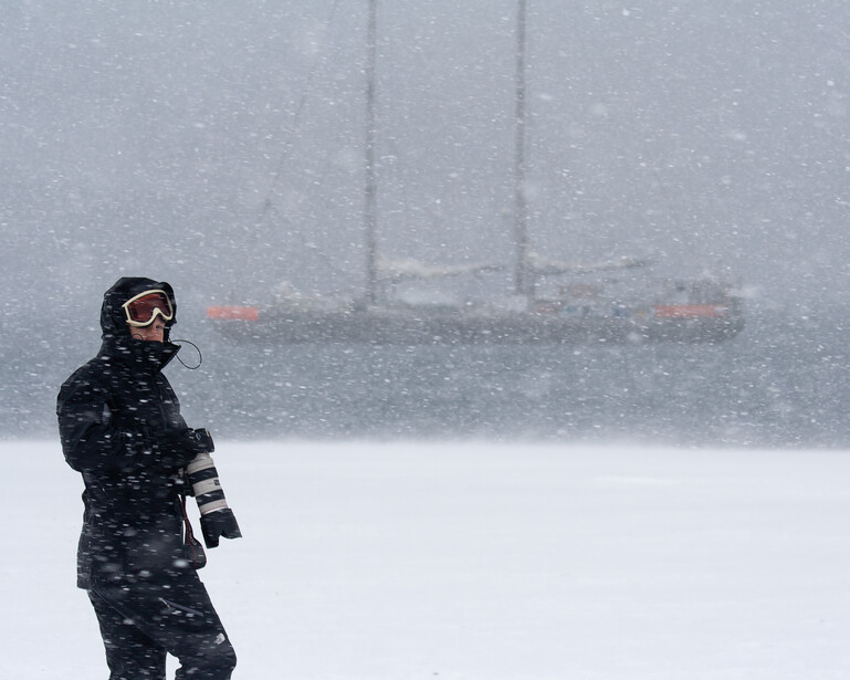 Snowboarder in winter gear standing on snowy terrain during heavy snowfall with vehicles visible in foggy background.