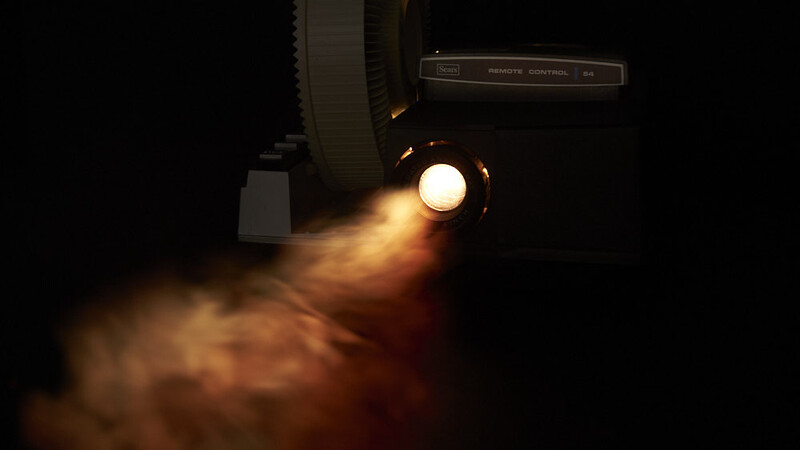 Vintage film projector with warm yellow light beam casting through dust and smoke against black background.