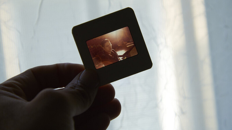 Hand holding a photographic slide up to bright light, revealing a warm-toned image of a person and glass on the film.