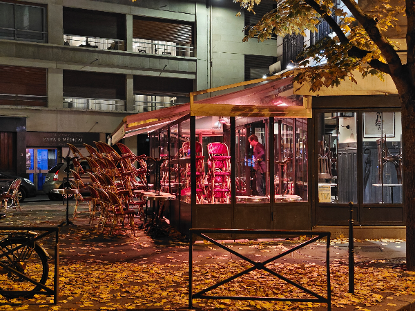 Red-lit kiosk or small storefront in urban plaza at dusk with autumn foliage overhead.