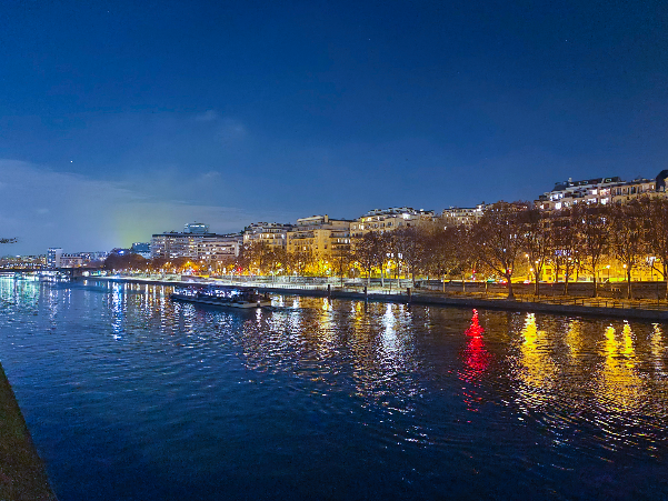Riverside city waterfront at dusk with illuminated buildings and a red navigation light reflecting on calm water.