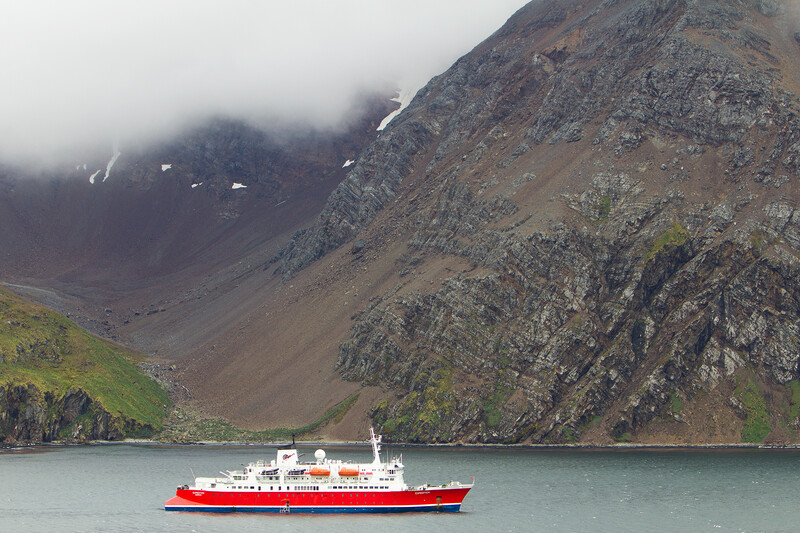 Red and white ship anchored in a fjord beneath steep, misty mountains with waterfalls.