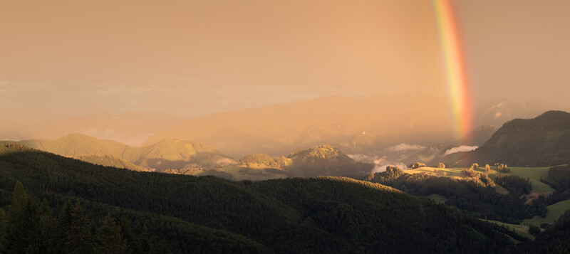 Rainbow extending from dark storm clouds over a forested valley with snow-capped mountains and golden sunlight breaking through clouds.